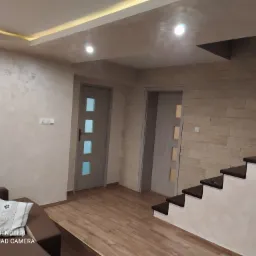 Nowoczesne wnętrze z dekoracyjną ścianą z imitacji kamienia, drewnopodobną podłogą, dwiema parami drzwi wewnętrznych z przeszkleniami i częściowo widocznymi schodami.