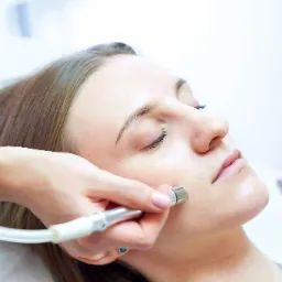 Kobieta podczas zabiegu mikrodermabrazji diamentowej na policzku, widoczne urządzenie z metalową końcówką i biały przewód, dłoń kosmetyczki z delikatnym różowym manicure.