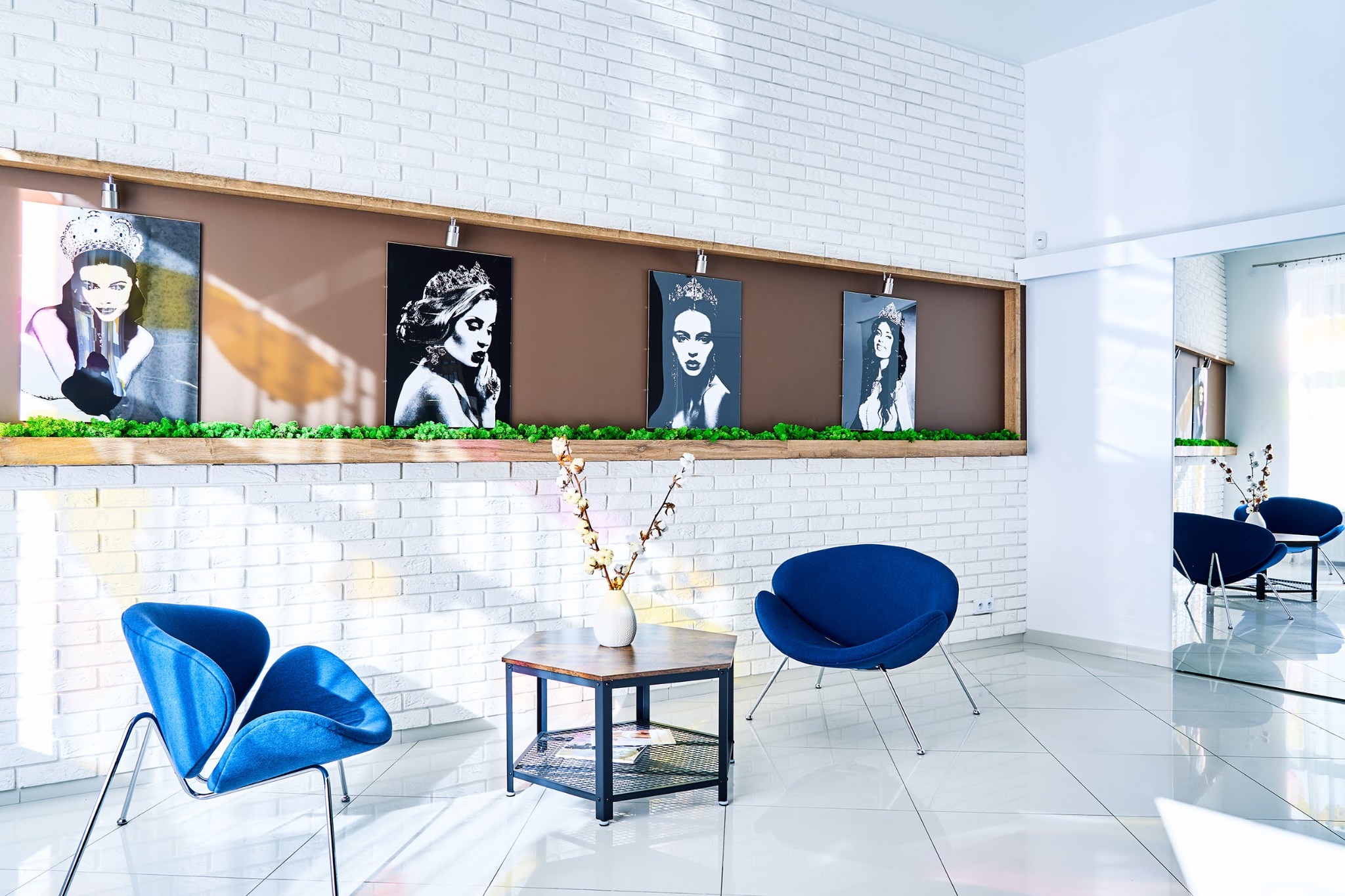 Jasne wnętrze salonu z białej cegły z dekoracjami ściennymi przedstawiającymi portrety kobiet w koronach, niebieskie designerskie fotele i stolik kawowy z wazonem z gałązkami bawełny.