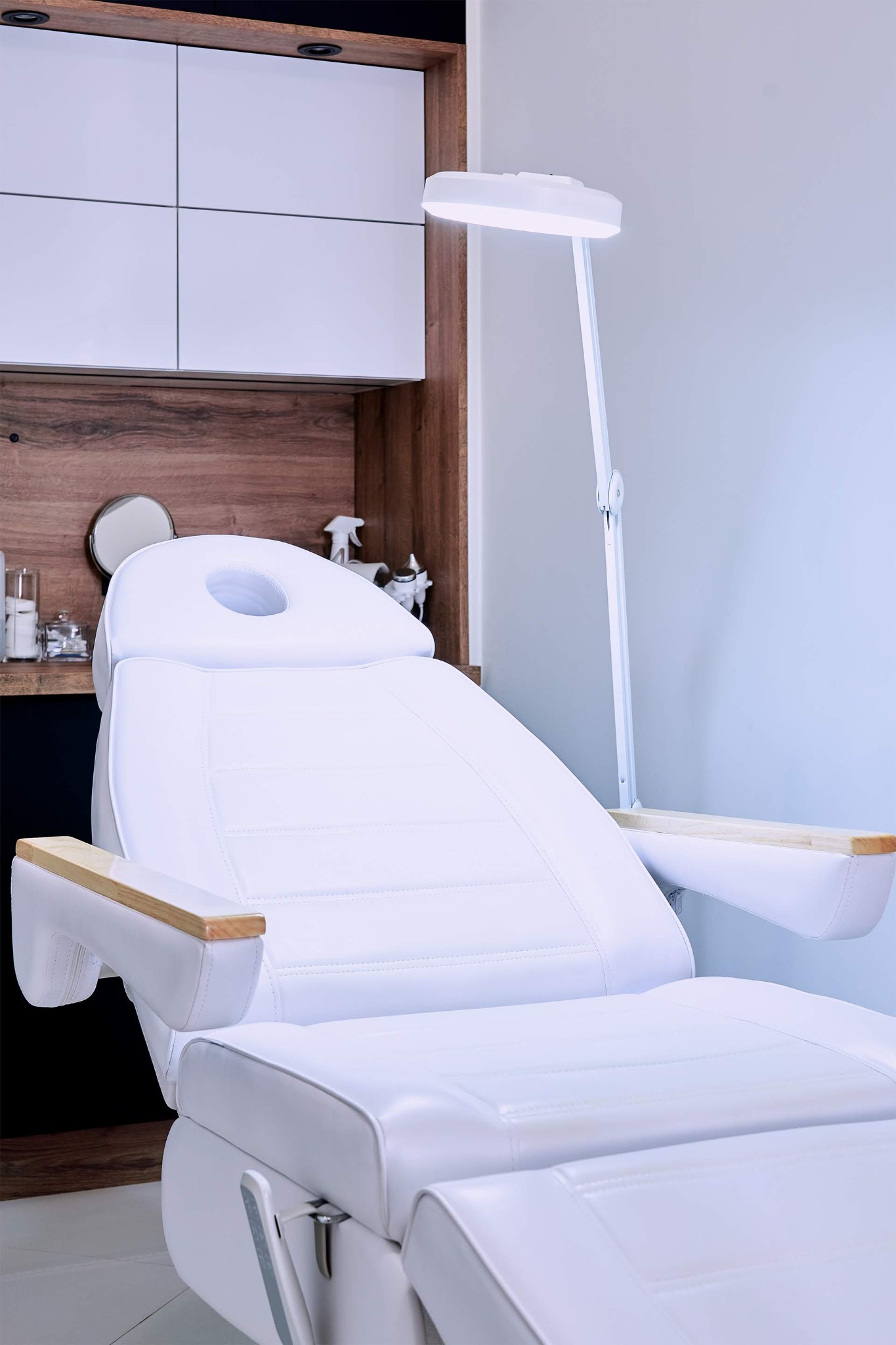 Białe, skórzane, regulowane łóżko do zabiegów kosmetycznych z drewnianymi podłokietnikami i lampą powiększającą w tle szafki z kosmetykami.