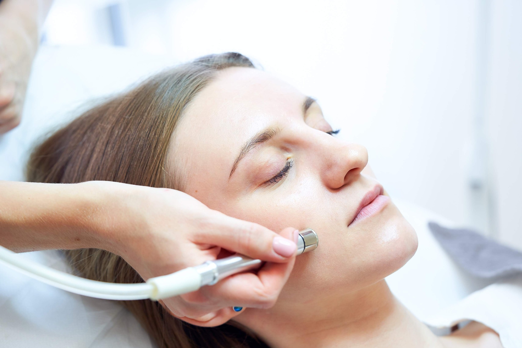 Kobieta podczas zabiegu mikrodermabrazji diamentowej na policzku, widoczne urządzenie z metalową końcówką i biały przewód, dłoń kosmetyczki z delikatnym różowym manicure.