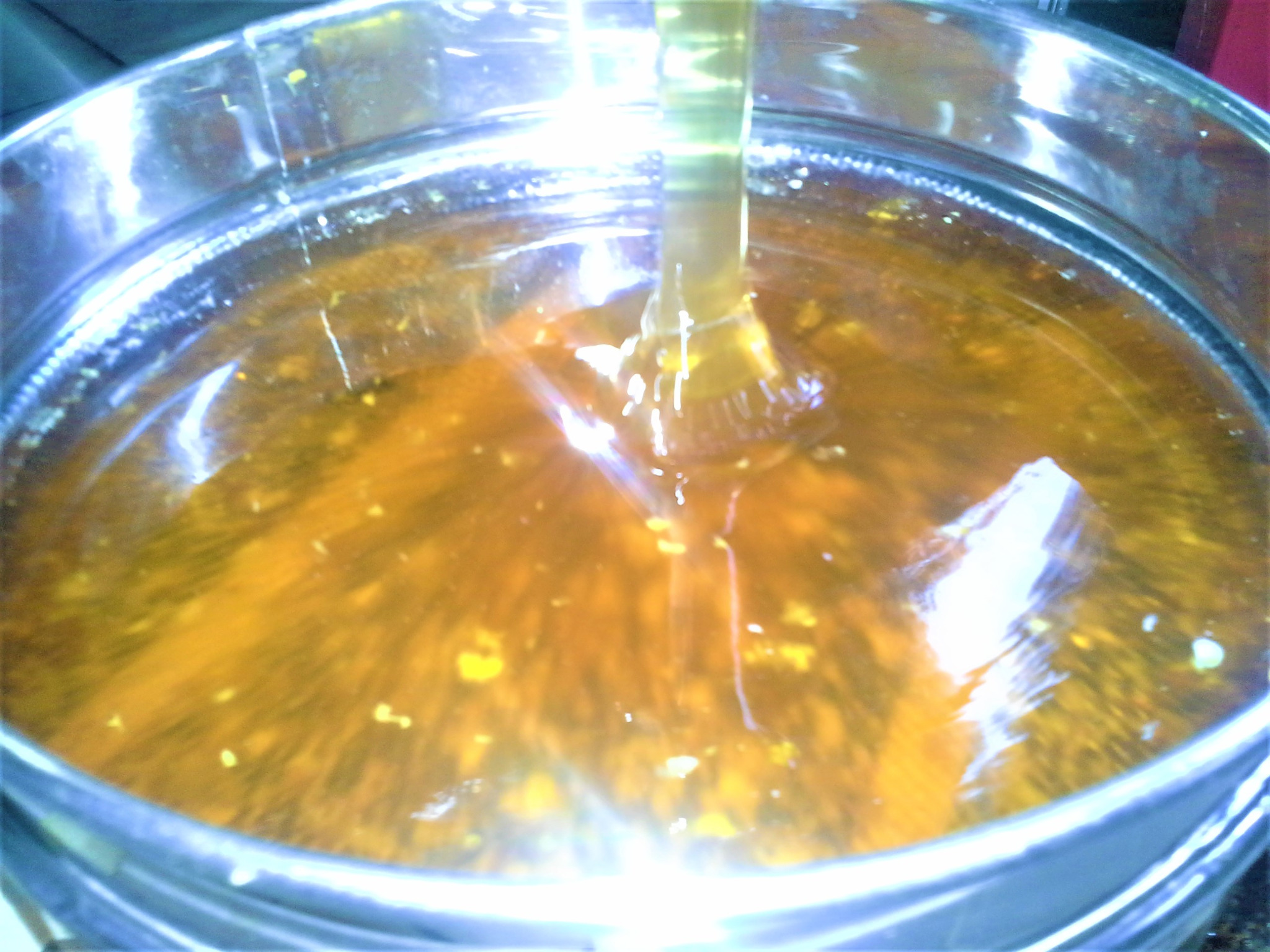 Strumień bursztynowego płynu wlewany do metalowego pojemnika, widoczne drobinki w zawiesinie.