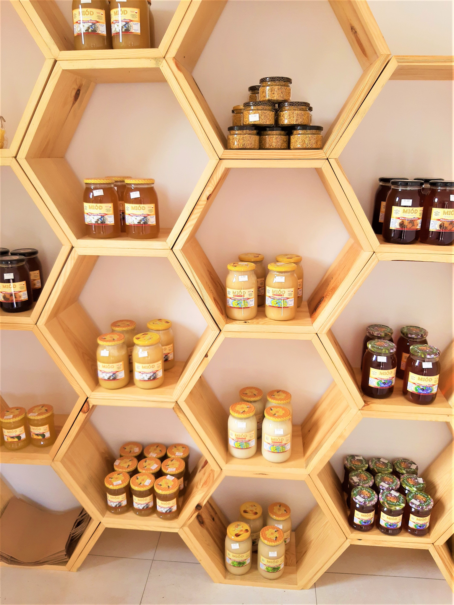 Półki w kształcie plastra miodu wypełnione słoikami z różnymi rodzajami miodu, pyłku pszczelego i miodów smakowych z owocami.