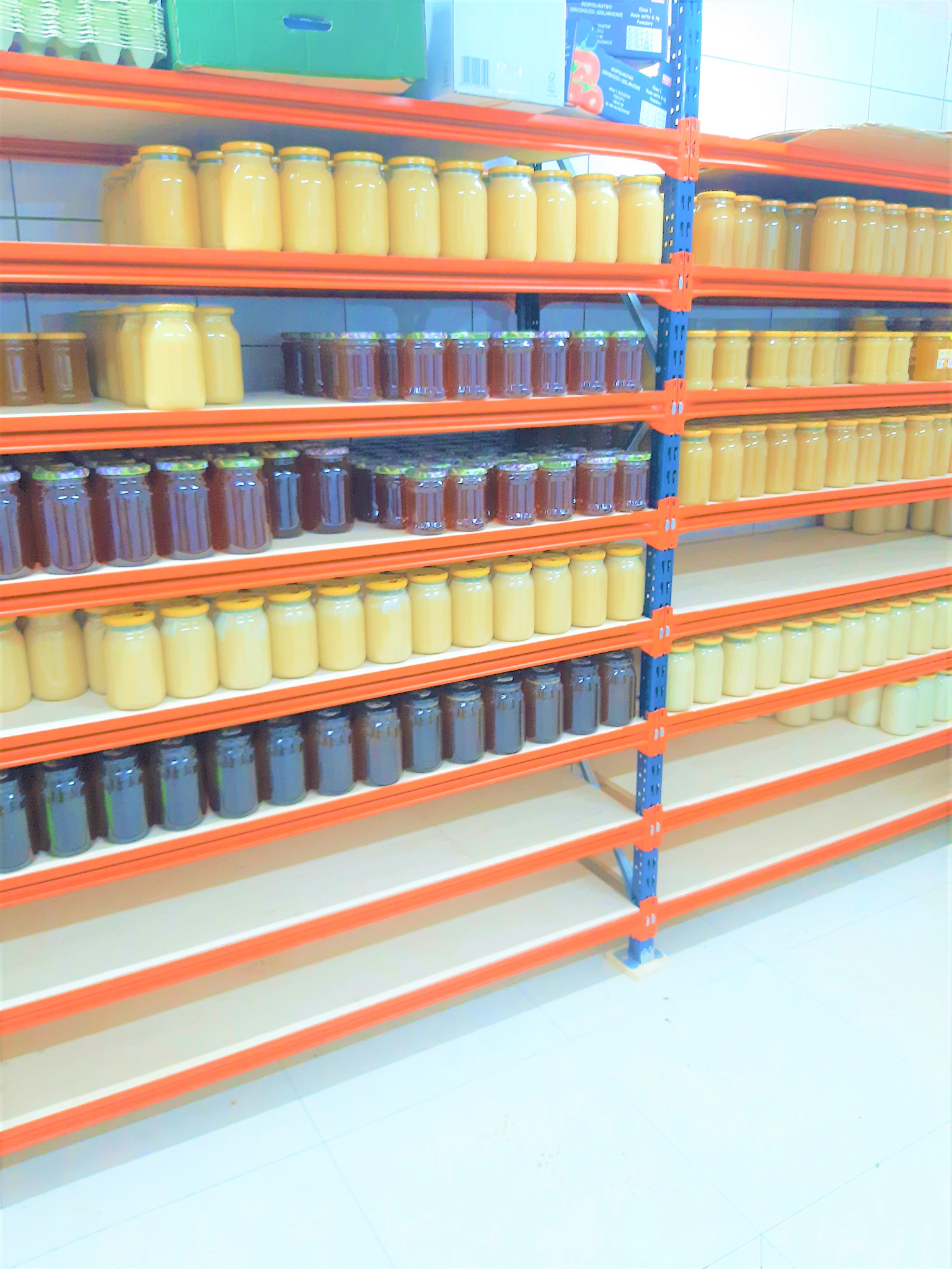 Magazyn z półkami wypełnionymi słoikami z różnymi rodzajami miodu, od jasnych po ciemne, ułożonymi rzędami na pomarańczowych regałach.