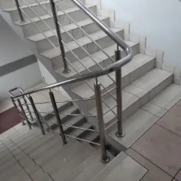 Schody wyłożone jasnymi płytkami z metalową balustradą z wypełnieniem z prętów, widok z góry, fragment podestu z brązowymi płytkami w tle.