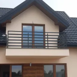 Nowoczesna, pozioma balustrada balkonowa z ciemnego metalu na tle beżowej elewacji domu z ciemną dachówką, widok z dołu.