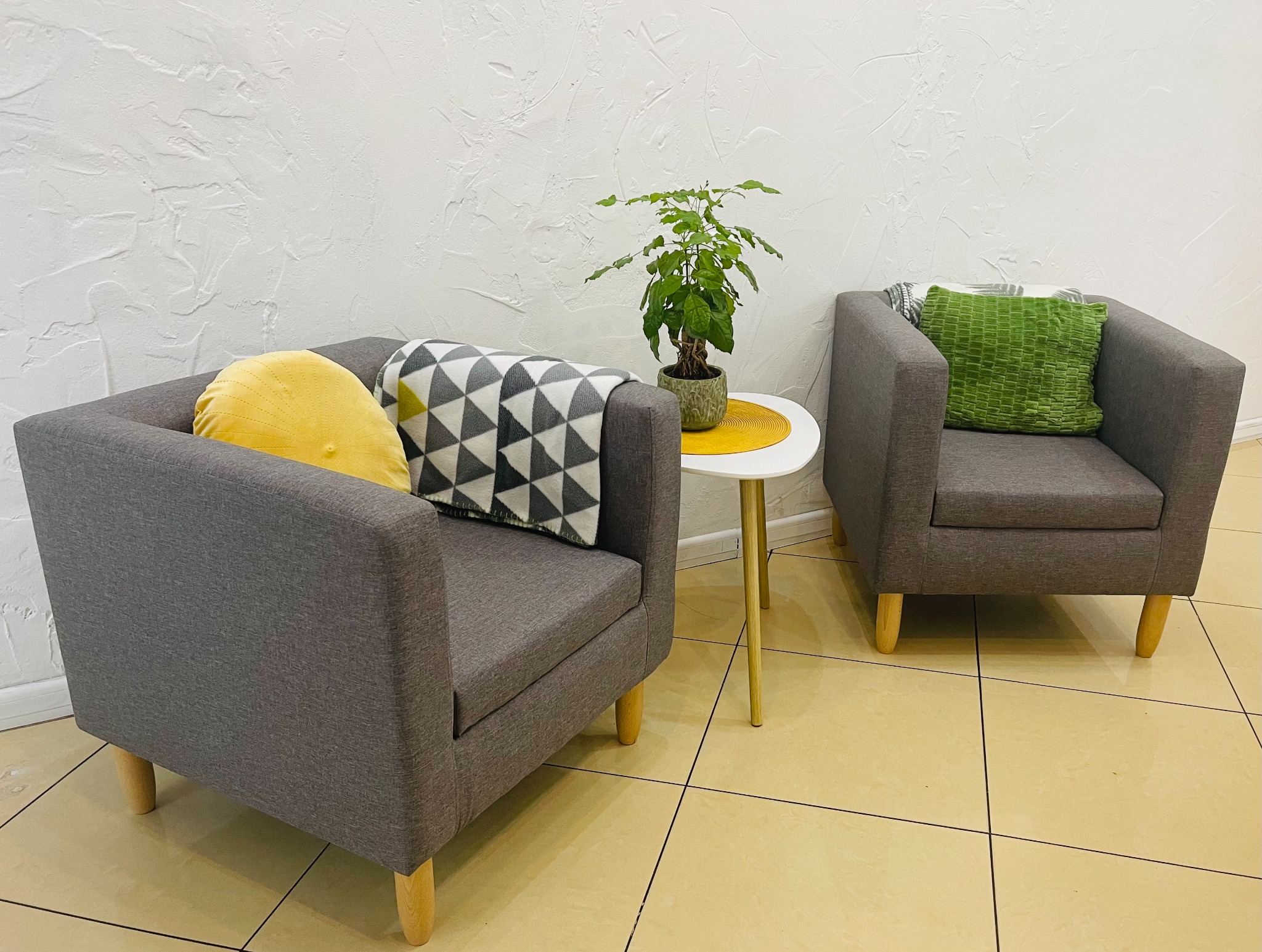 Dwa szare fotele z drewnianymi nogami, jeden z żółtą okrągłą poduszką i kocem w szare trójkąty, drugi z zieloną kwadratową poduszką, pomiędzy nimi stolik z rośliną w doniczce na żółtej podkładce...