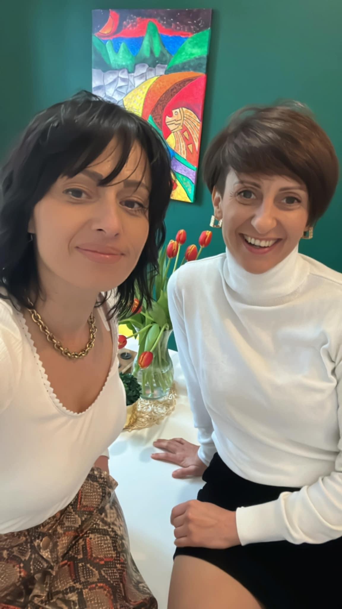 Portret dwóch uśmiechniętych kobiet w jasnych ubraniach, z wazonem tulipanów i abstrakcyjnym obrazem w tle.