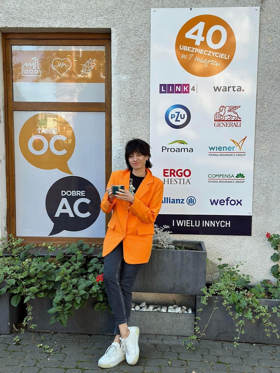Kobieta w pomarańczowej marynarce pozuje z kubkiem na tle szyldu z logo firm ubezpieczeniowych i okna z napisem 'Tanie OC' i 'Dobre AC'