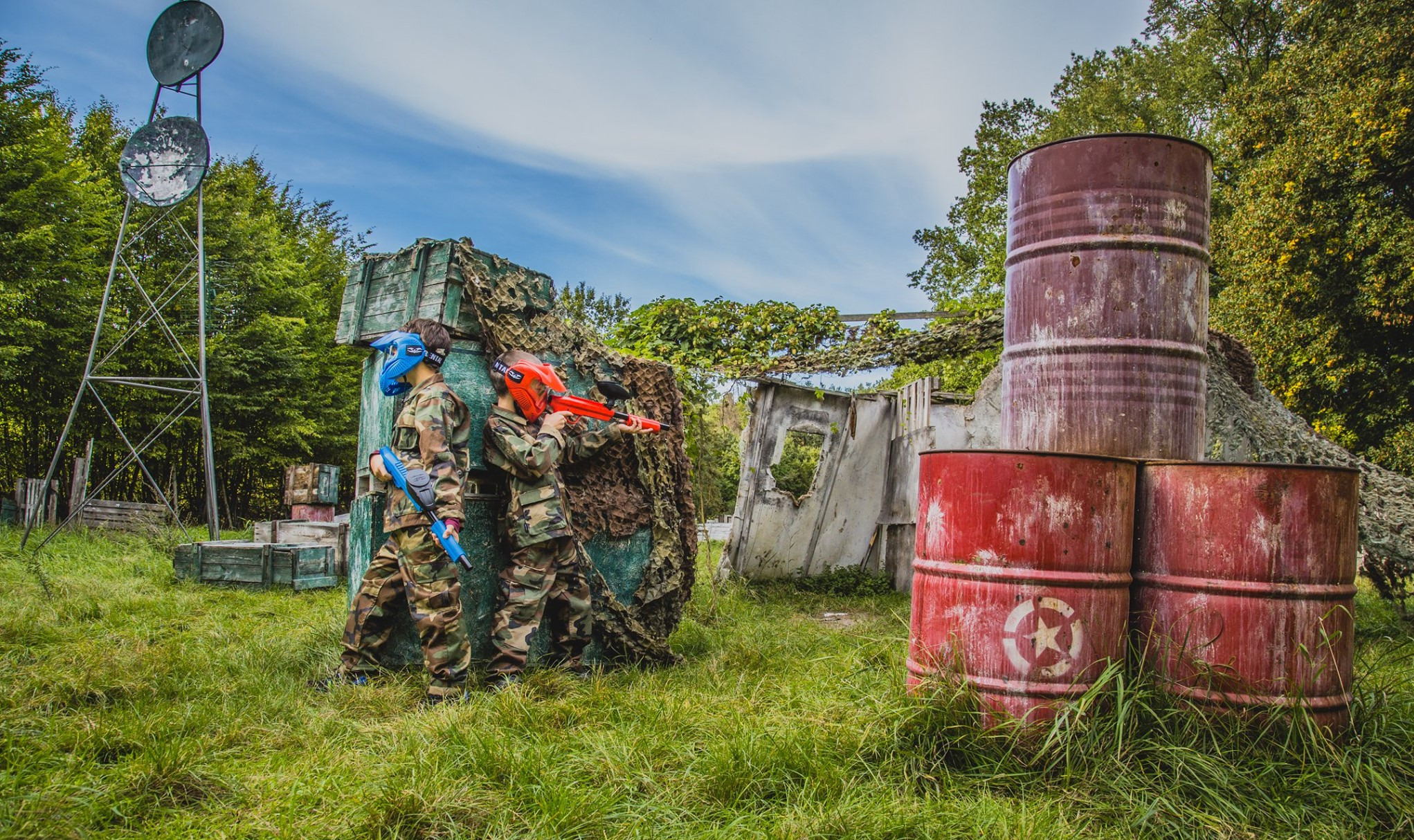 Dwoje dzieci w strojach kamuflażowych i maskach ochronnych celuje z pistoletów do paintballa w scenerii z beczek i siatek maskujących na terenie gry.
