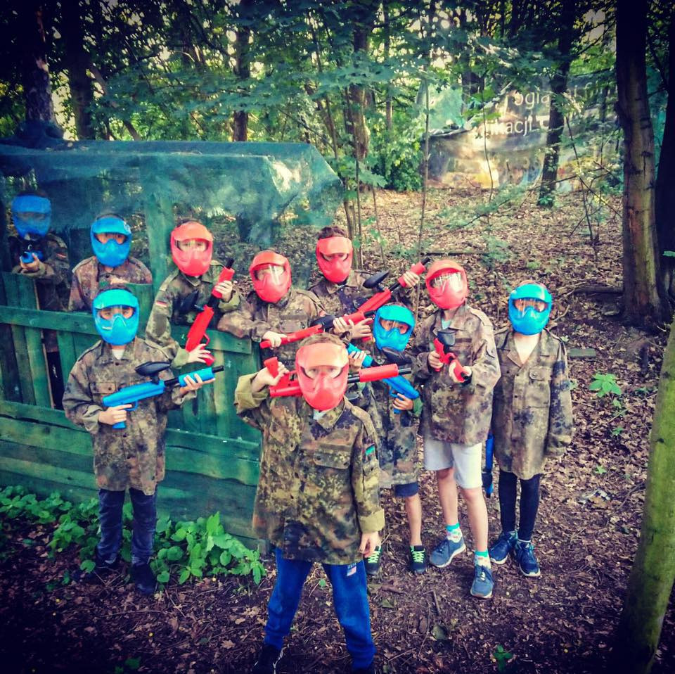 Grupa dzieci w maskach i strojach moro pozuje z pistoletami do paintballa na tle leśnej scenerii z drewnianą konstrukcją.