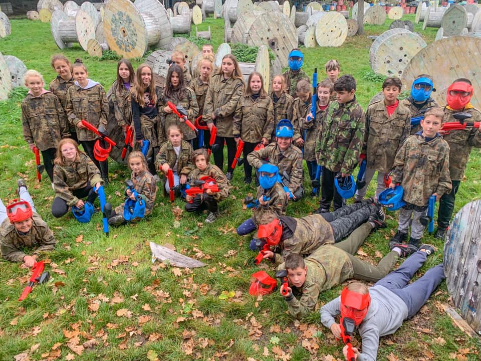 Grupa dzieci w strojach moro i maskach ochronnych, trzymających markery paintballowe, pozuje na trawiastym terenie z drewnianymi szpulami w tle.