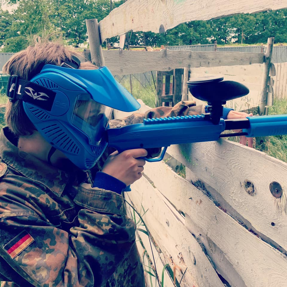 Osoba w kamuflażu i niebieskiej masce ochronnej celuje z niebieskiego markera paintballowego zza drewnianej przeszkody z niemiecką flagą na ramieniu.