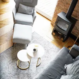 Jasne wnętrze z drewnianą podłogą w jodełkę, szarym fotelem z podnóżkiem, dwoma okrągłymi stolikami kawowymi na wzorzystym dywanie oraz czarnym piecem wolnostojącym z rurą kominową przy ceglanej...