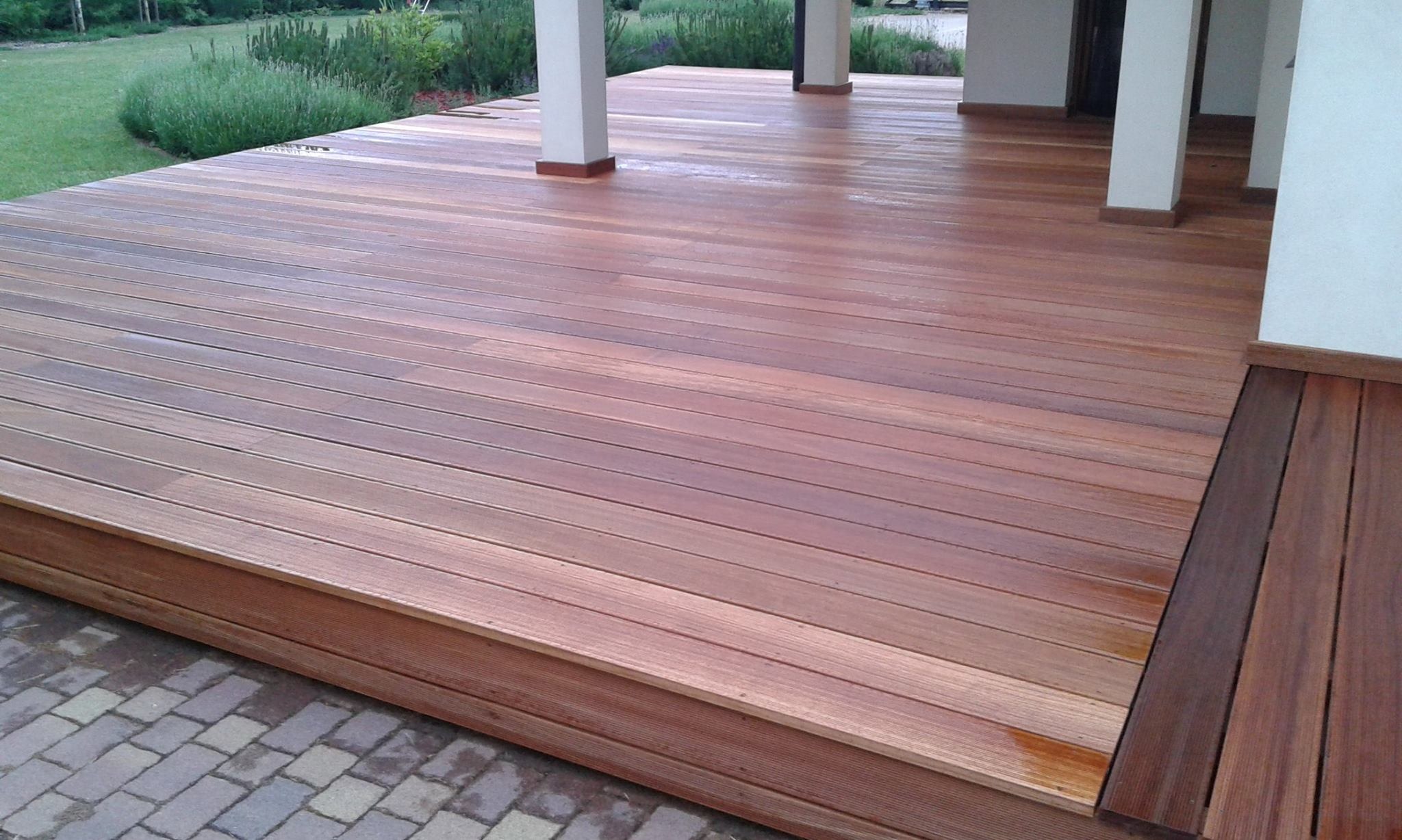 Nowoczesny taras z desek o ciepłym odcieniu drewna, z niskim stopniem wejściowym i widocznymi białymi kolumnami podtrzymującymi zadaszenie, otoczony zielenią ogrodu.