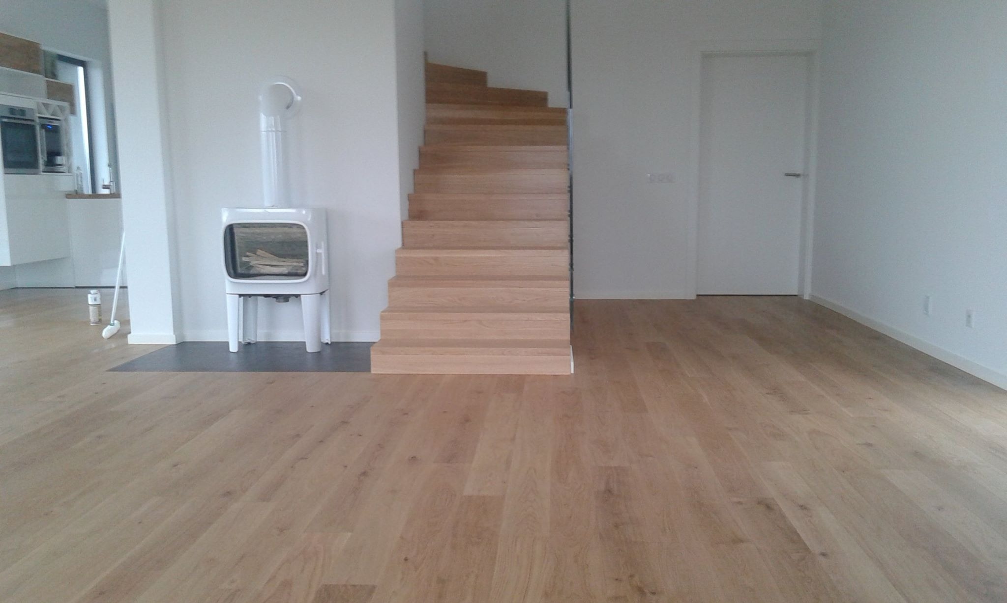 Jasna podłoga drewniana w minimalistycznym wnętrzu z białym piecem wolnostojącym i drewnianymi schodami bez poręczy.