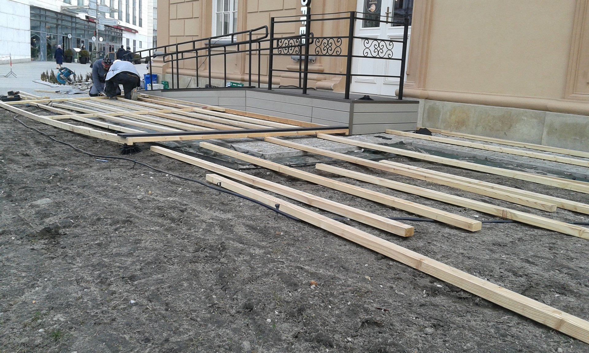 Montaż drewnianej konstrukcji pod taras przy budynku z rampą dla niepełnosprawnych, widoczna podbudowa z legarów i desek, dwóch pracowników w trakcie pracy.