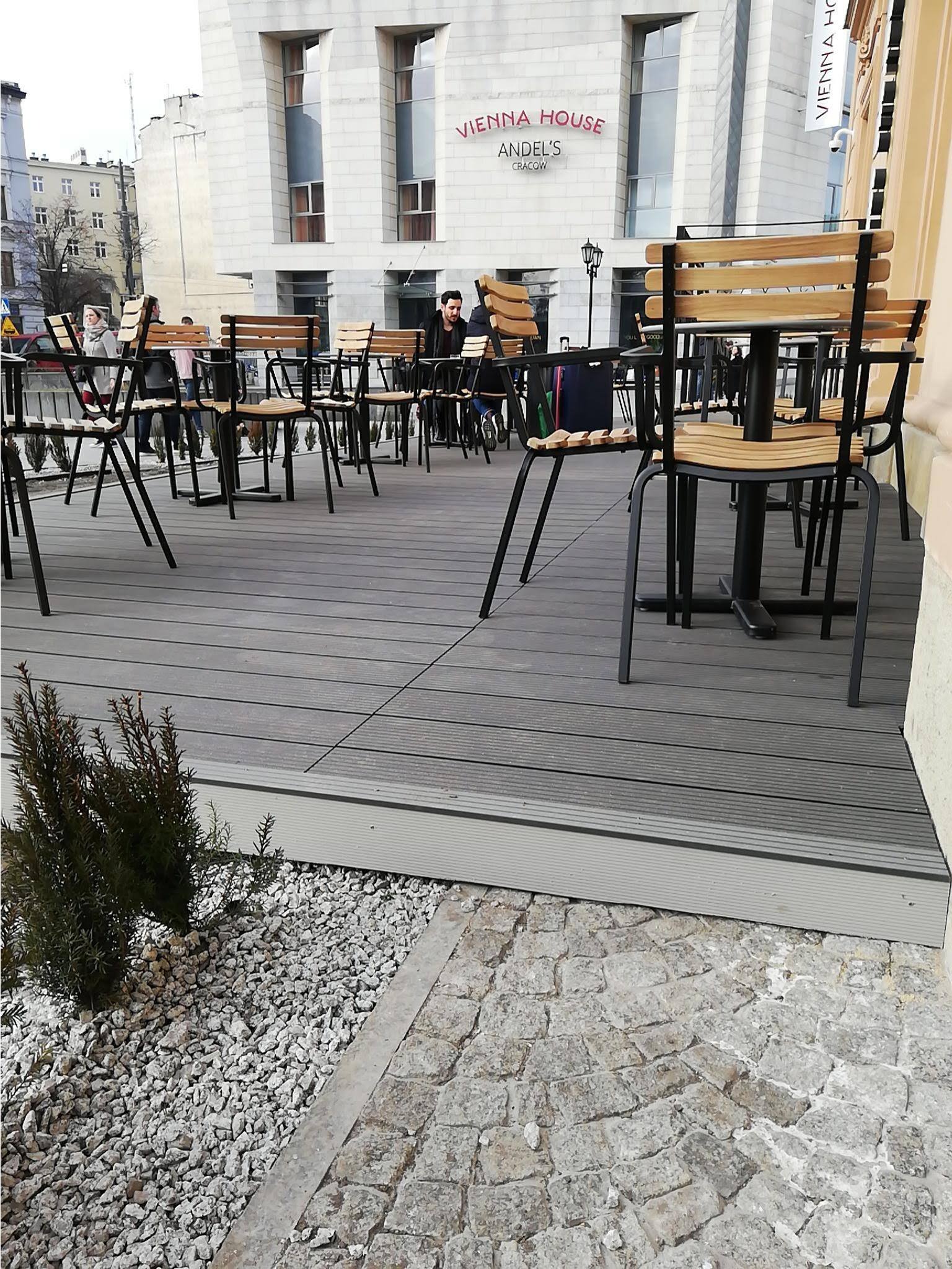 Zewnętrzny taras restauracji z szarej deski kompozytowej, widoczne stoliki z krzesłami i ogródek z kamieniami.