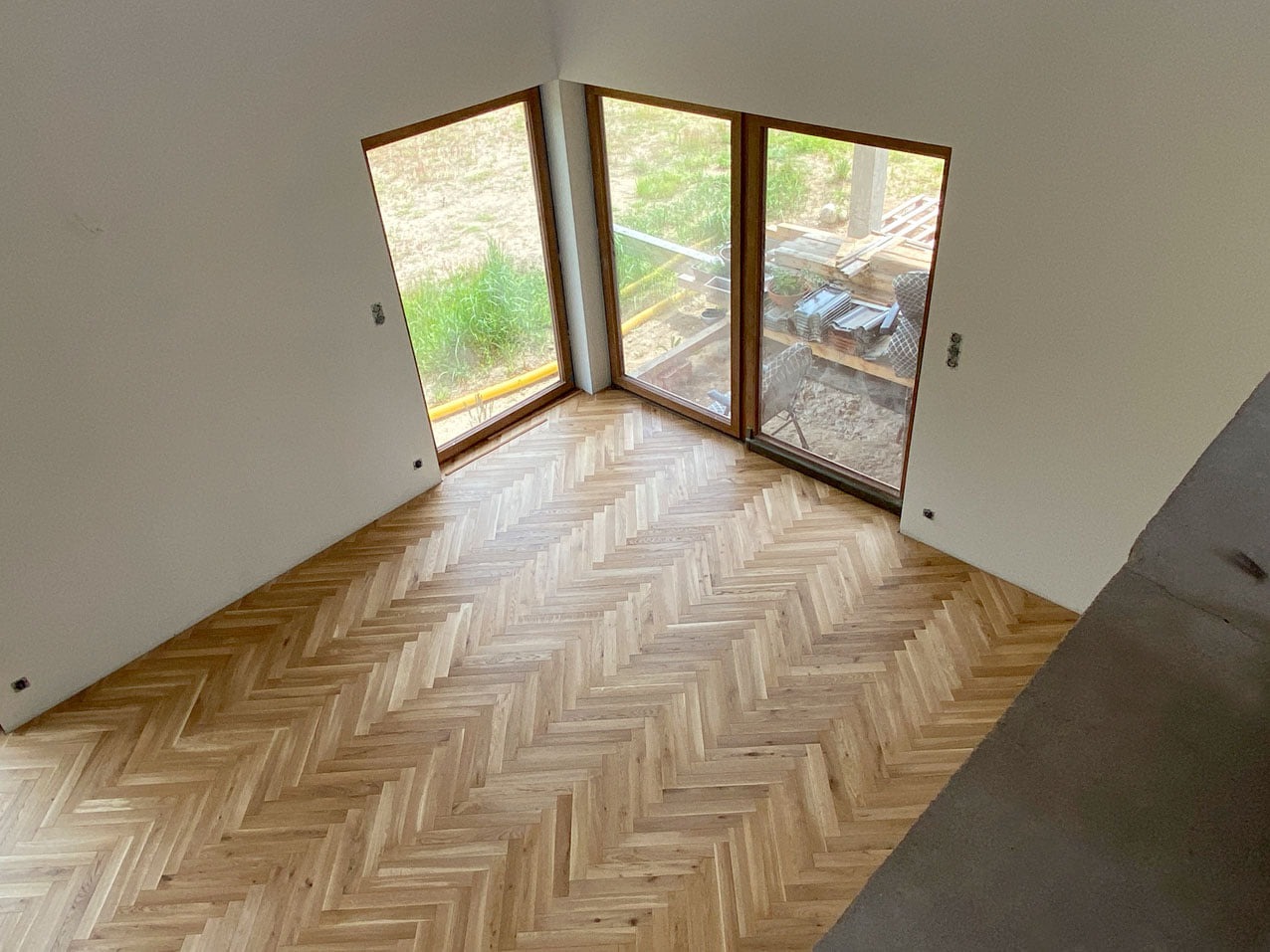 Wnętrze z nową, jasną podłogą drewnianą ułożoną w jodełkę, widok z góry na dwa okna wychodzące na ogród, na zewnątrz widać materiały budowlane.