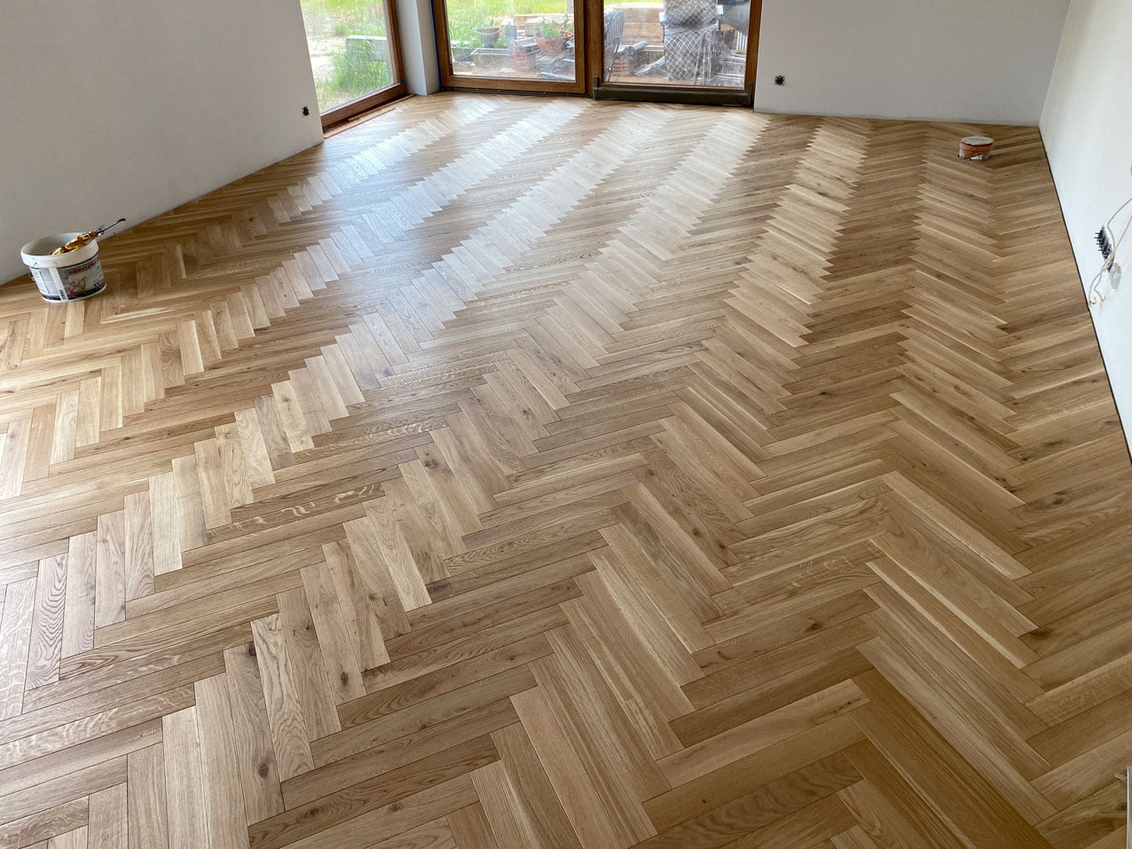 Ułożona w jodełkę, jasna podłoga drewniana w nowym pomieszczeniu, z widocznymi oknami i wiaderkiem z farbą w rogu, sugerującym trwające prace wykończeniowe.