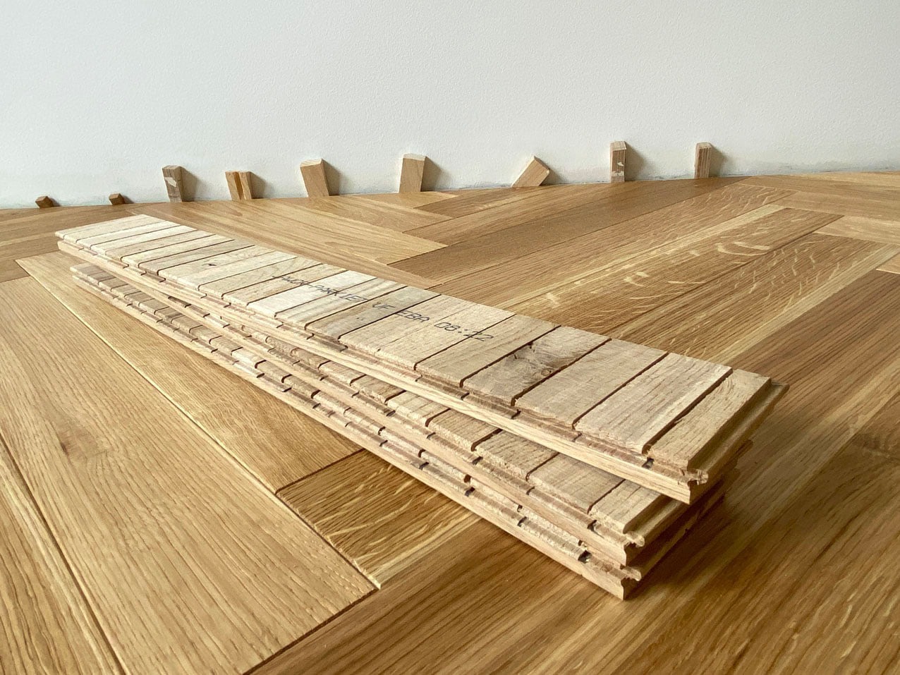 Układanie parkietu dębowego w jodełkę, widoczne paczki desek i kliny dystansowe przy ścianie.