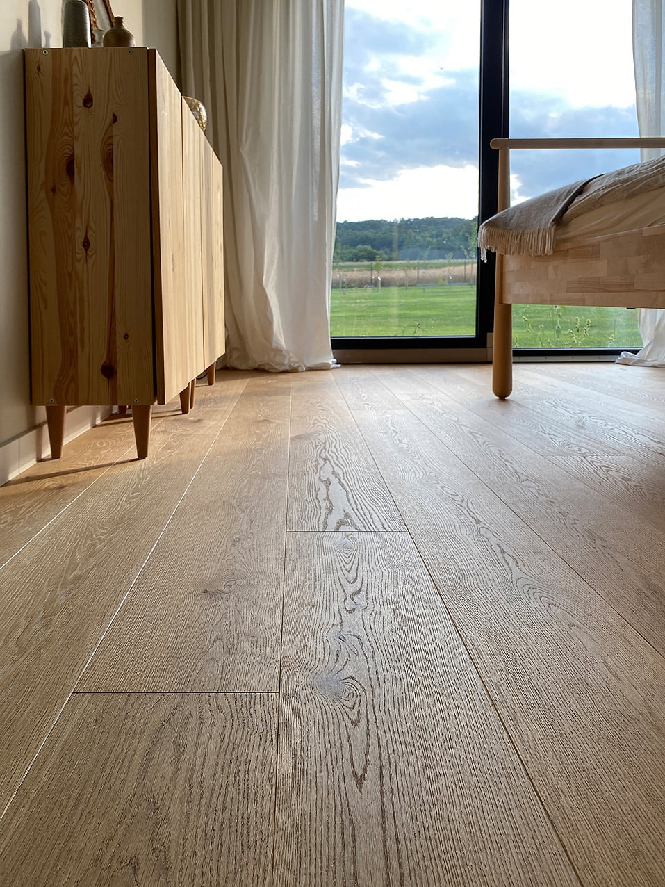 Jasne, drewniane deski podłogowe ułożone w salonie z widokiem na zielony krajobraz za oknem, widoczna szafka z jasnego drewna i fragment łóżka.