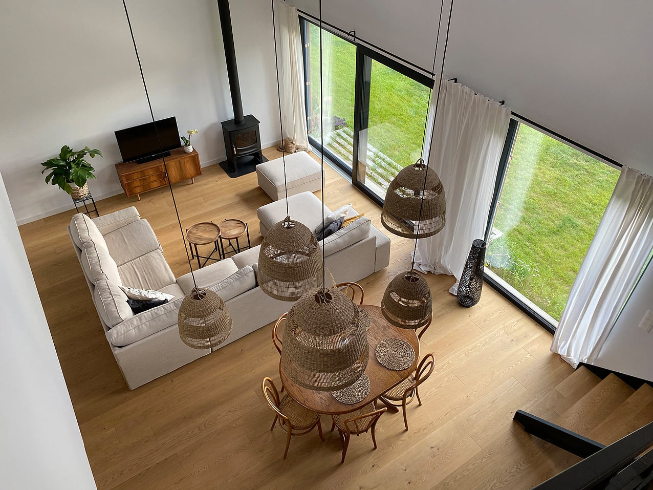 Wnętrze domu z drewnianą podłogą, widok z góry, z dużymi oknami wychodzącymi na trawnik, oświetlone wiszącymi lampami z abażurami z wikliny.