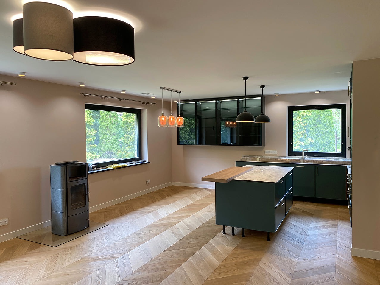 Jasny salon z podłogą ułożoną w jodełkę, nowoczesnym piecem wolnostojącym na szklanej podstawie, zieloną zabudową kuchenną i wyspą z drewnianym blatem.