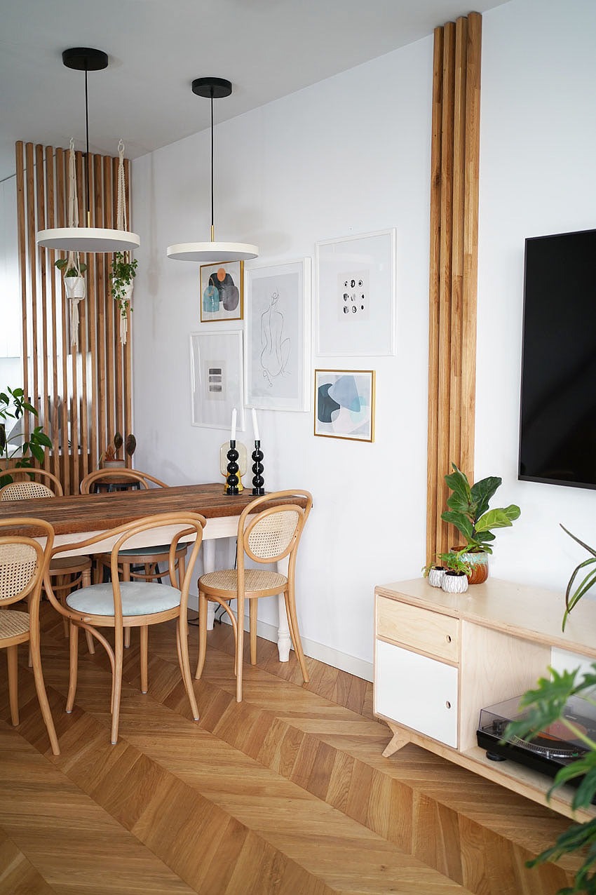 Jasna jadalnia z parkietem ułożonym w jodełkę, drewnianymi krzesłami z rattanowym oparciem, stołem z drewnianym blatem i białymi nogami, oraz ścianą z listew drewnianych i dekoracjami.