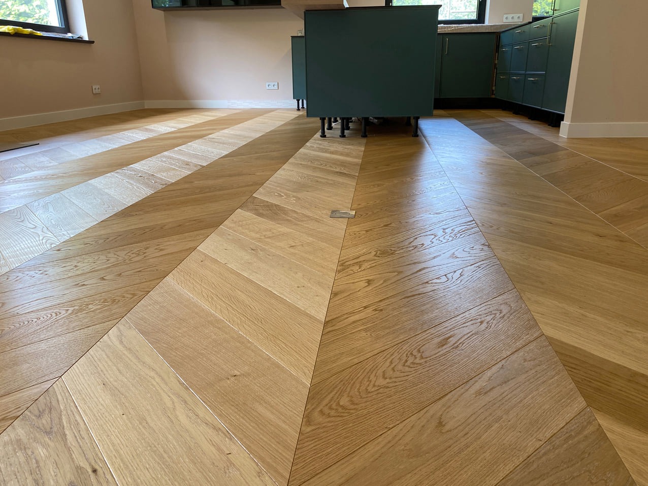 Parkiet ułożony we wzór jodełki francuskiej w nowoczesnej kuchni z zielonymi szafkami, widoczny fragment podłogi z różnicą w odcieniach drewna.