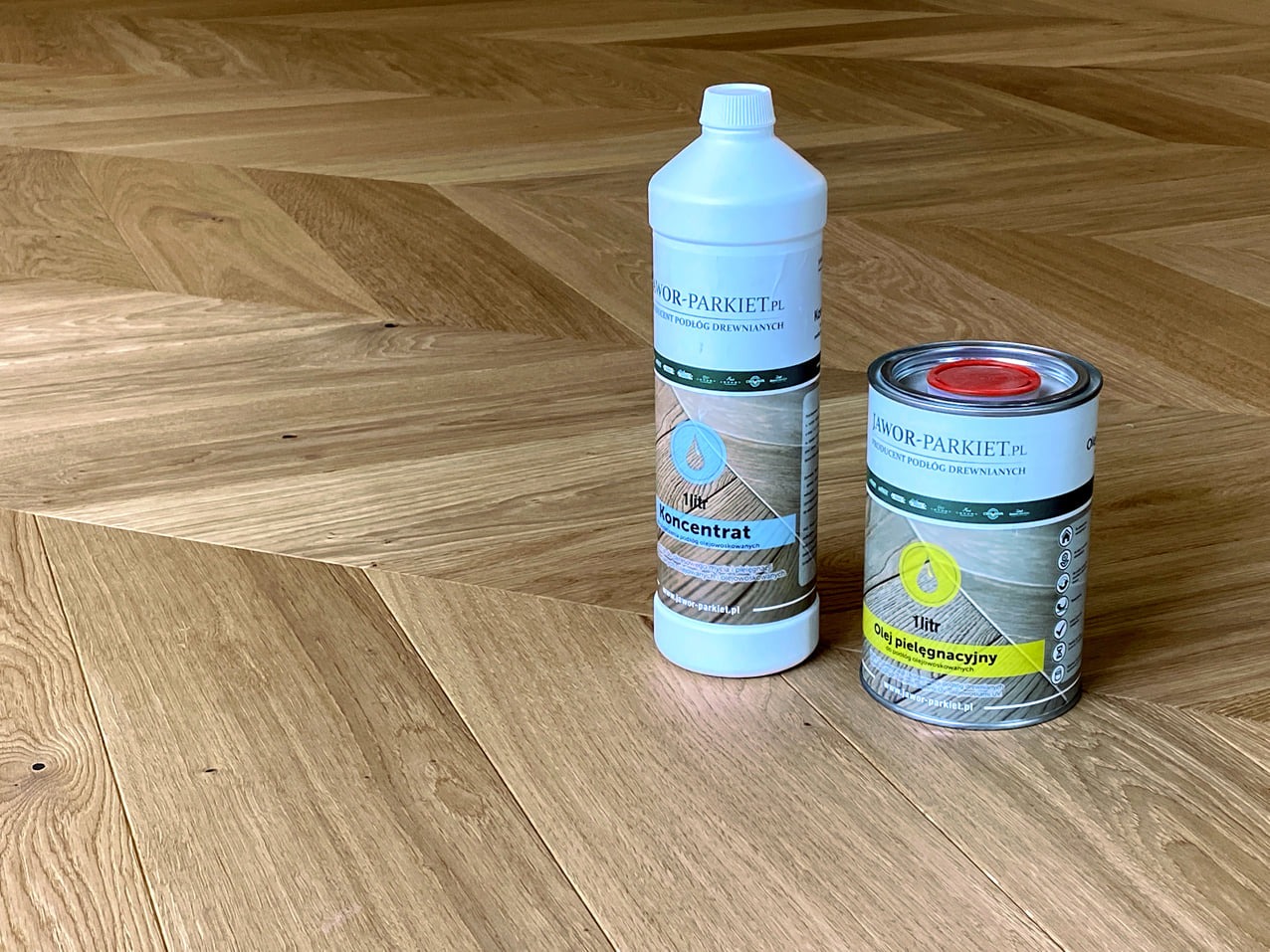 Parkiet ułożony we wzór jodełki z butelką koncentratu i puszką oleju pielęgnacyjnego do drewna firmy Jawor-Parkiet.