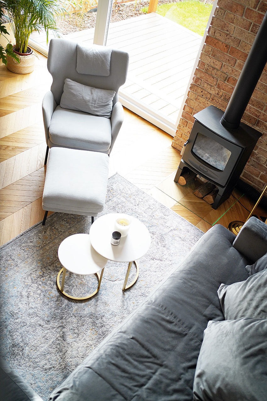 Jasne wnętrze z drewnianą podłogą w jodełkę, szarym fotelem z podnóżkiem, dwoma okrągłymi stolikami kawowymi na wzorzystym dywanie oraz czarnym piecem wolnostojącym z rurą kominową przy ceglanej...