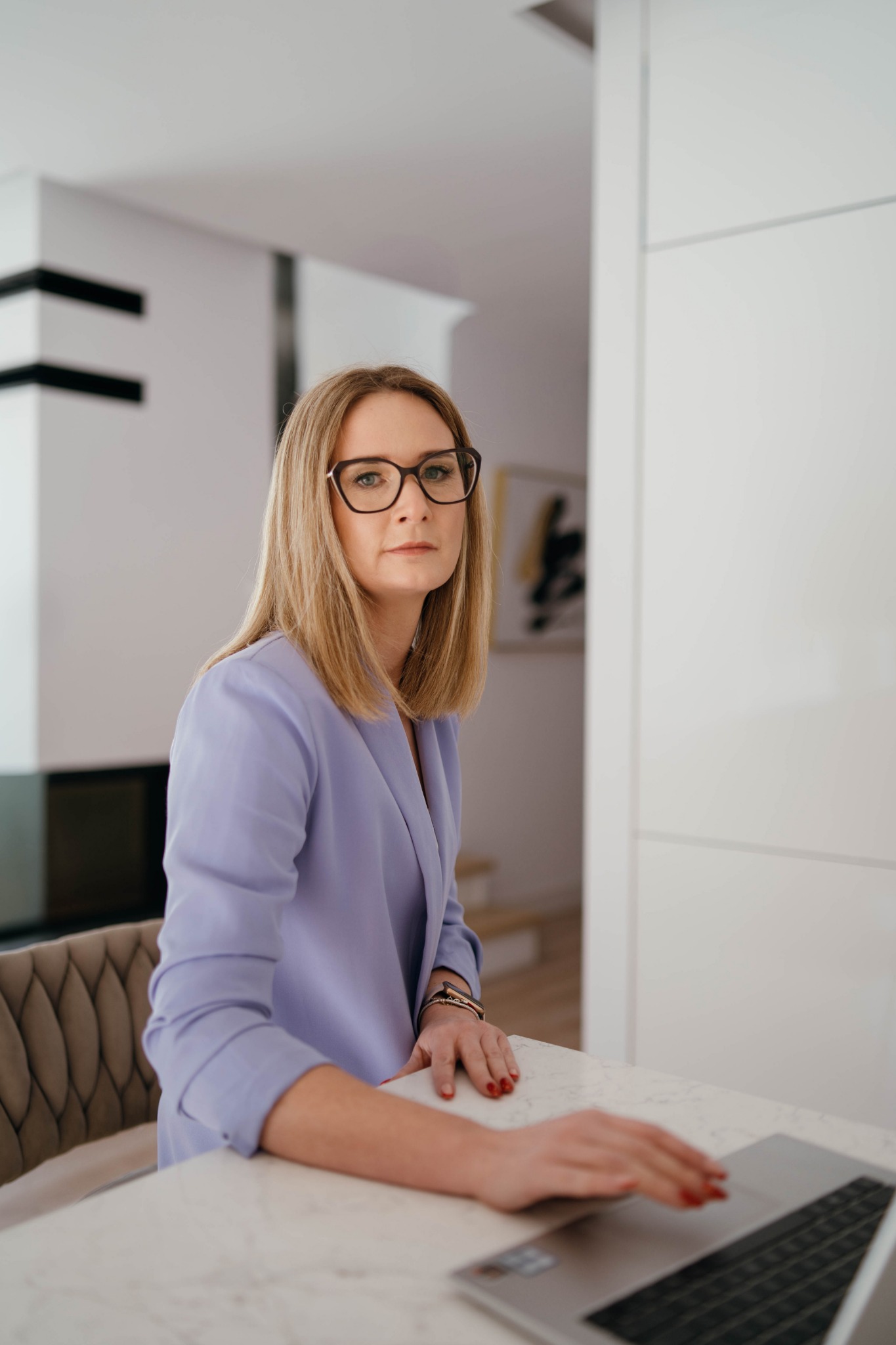 Kobieta w liliowej marynarce i okularach pracuje na laptopie przy marmurowym stole w nowoczesnym wnętrzu. Profesjonalny wizerunek.