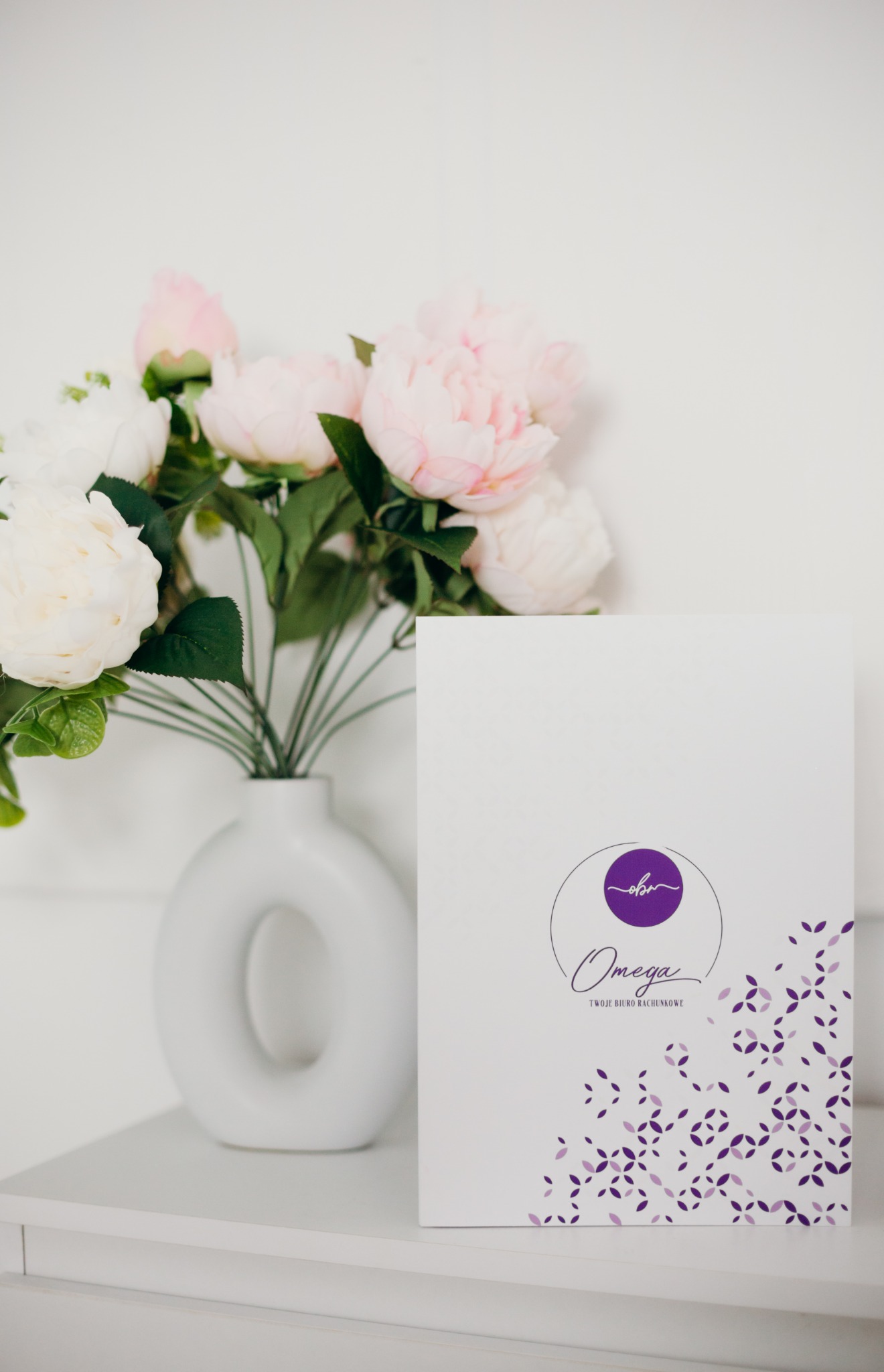 Wazon z pastelowymi kwiatami obok białej teczki z fioletowym logo 'Omega Twoje Biuro Rachunkowe' na białym tle.
