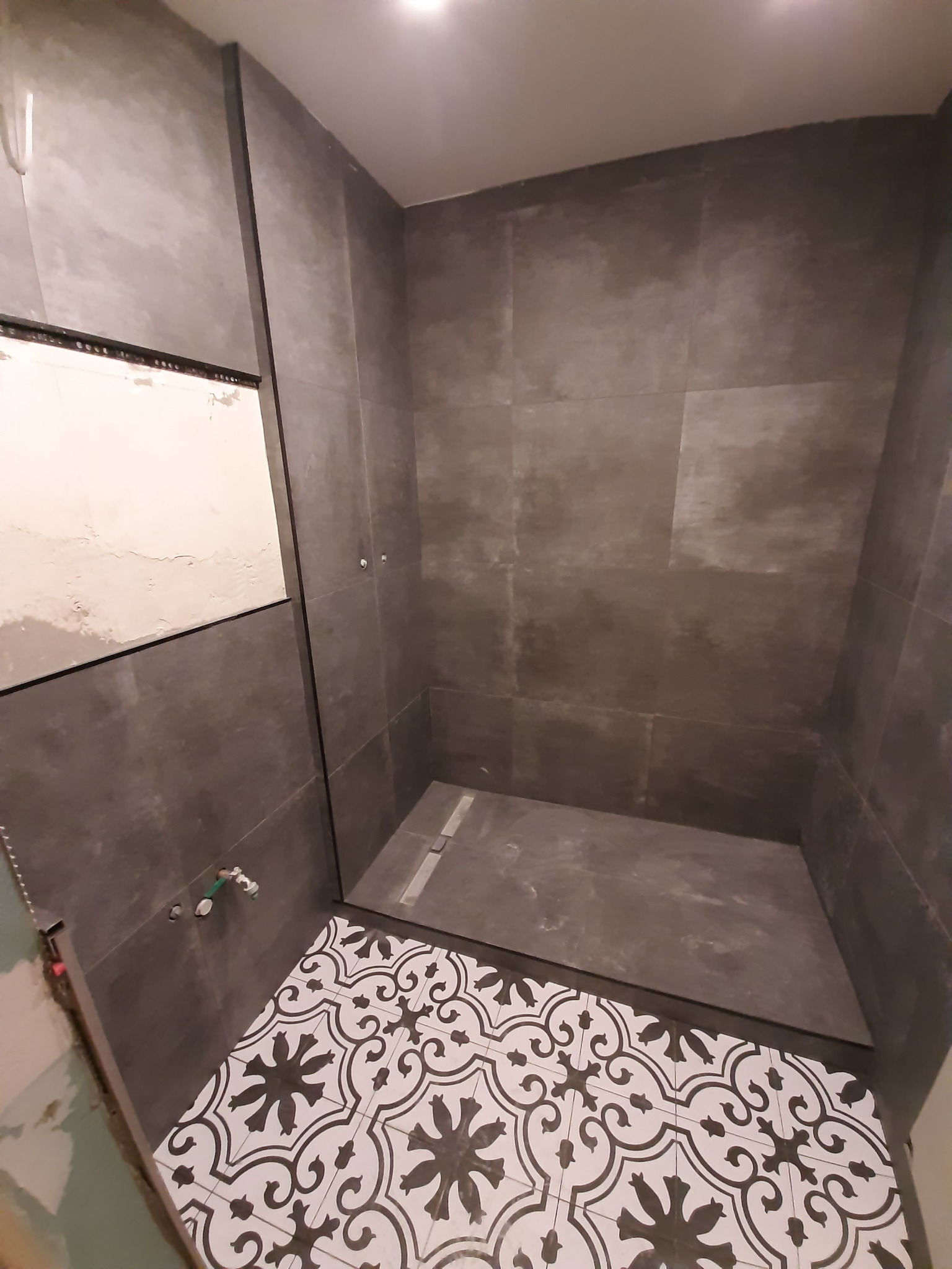 Łazienka w trakcie prac wykończeniowych: ściany wyłożone szarymi płytkami wielkoformatowymi, podłoga z biało-czarną wzorzystą mozaiką, widoczny odpływ liniowy w kabinie prysznicowej typu walk-in...