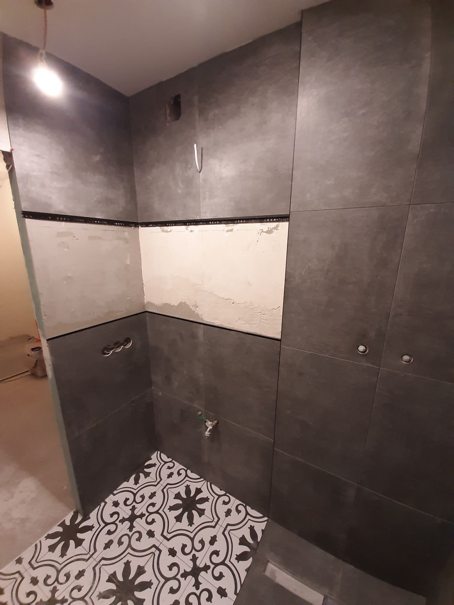 Łazienka w trakcie prac: ciemne płytki częściowo położone, fragment ściany przygotowany pod płytki, dekoracyjna podłoga z wzorem w gwiazdy i ornamenty.