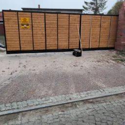 Zewnętrzna, przesuwna brama z drewnianym wypełnieniem i czarną, stalową ramą, z żółtą tabliczką informującą o monitoringu, obok skrzynka narzędziowa i oparta o bramę listwa.