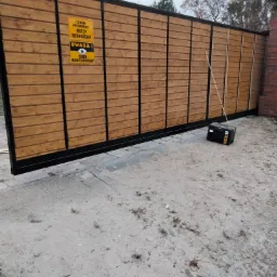 Przesuwna brama wjazdowa z drewnianym wypełnieniem i czarną ramą, tabliczki ostrzegawcze 'Teren prywatny' i skrzynka narzędziowa obok.