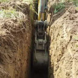Wąski wykop w ziemi wykonany koparką, widoczne ślady pracy łyżki, świeżo naruszona struktura gleby.