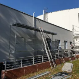 Szara elewacja budynku w trakcie malowania, widoczne rusztowania, drabina oparta o rusztowanie, pracownik w tle przy oknie, zabezpieczona podstawa budynku folią.
