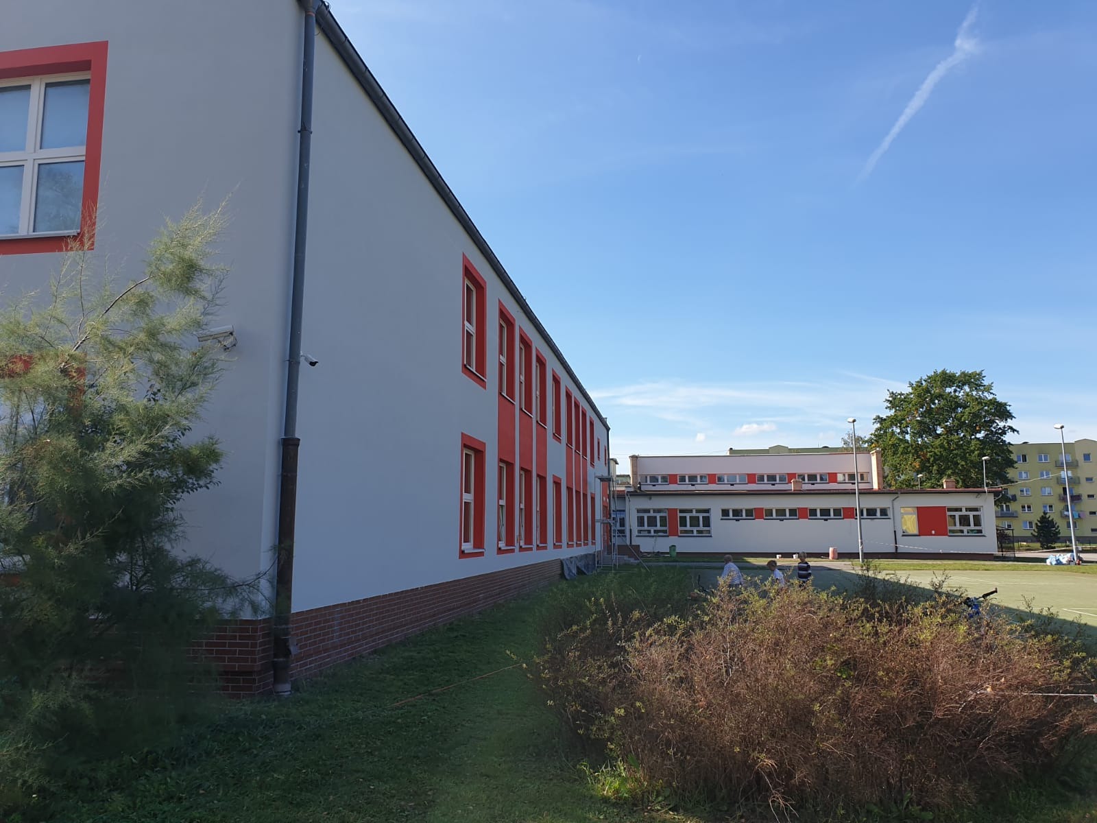 Odnowiona elewacja budynku szkolnego w odcieniach szarości z pomarańczowymi akcentami wokół okien, widoczne fragmenty muru z cegły, zieleń i niebieskie niebo w tle.