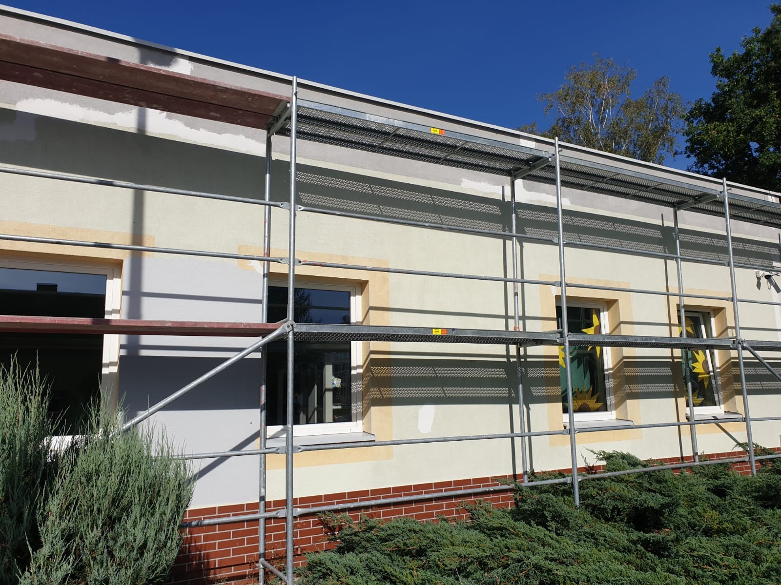Budynek z nałożonym tynkiem i przygotowany pod malowanie, widoczne rusztowanie, okna z żółtymi obramowaniami i częściowo ceglana podmurówka. Na oknach naklejone dekoracje w kształcie słońca.