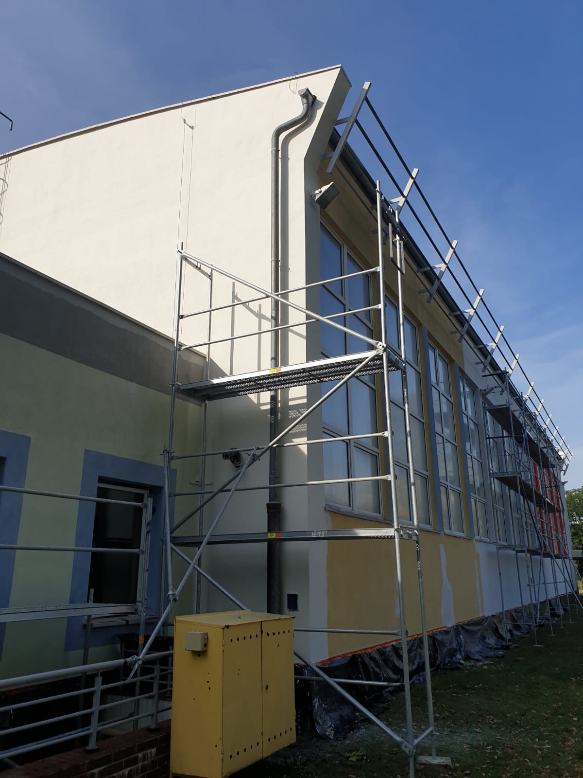Budynek w trakcie renowacji elewacji, widoczne rusztowania, częściowo pomalowana ściana, żółta skrzynka elektryczna przy ścianie budynku.