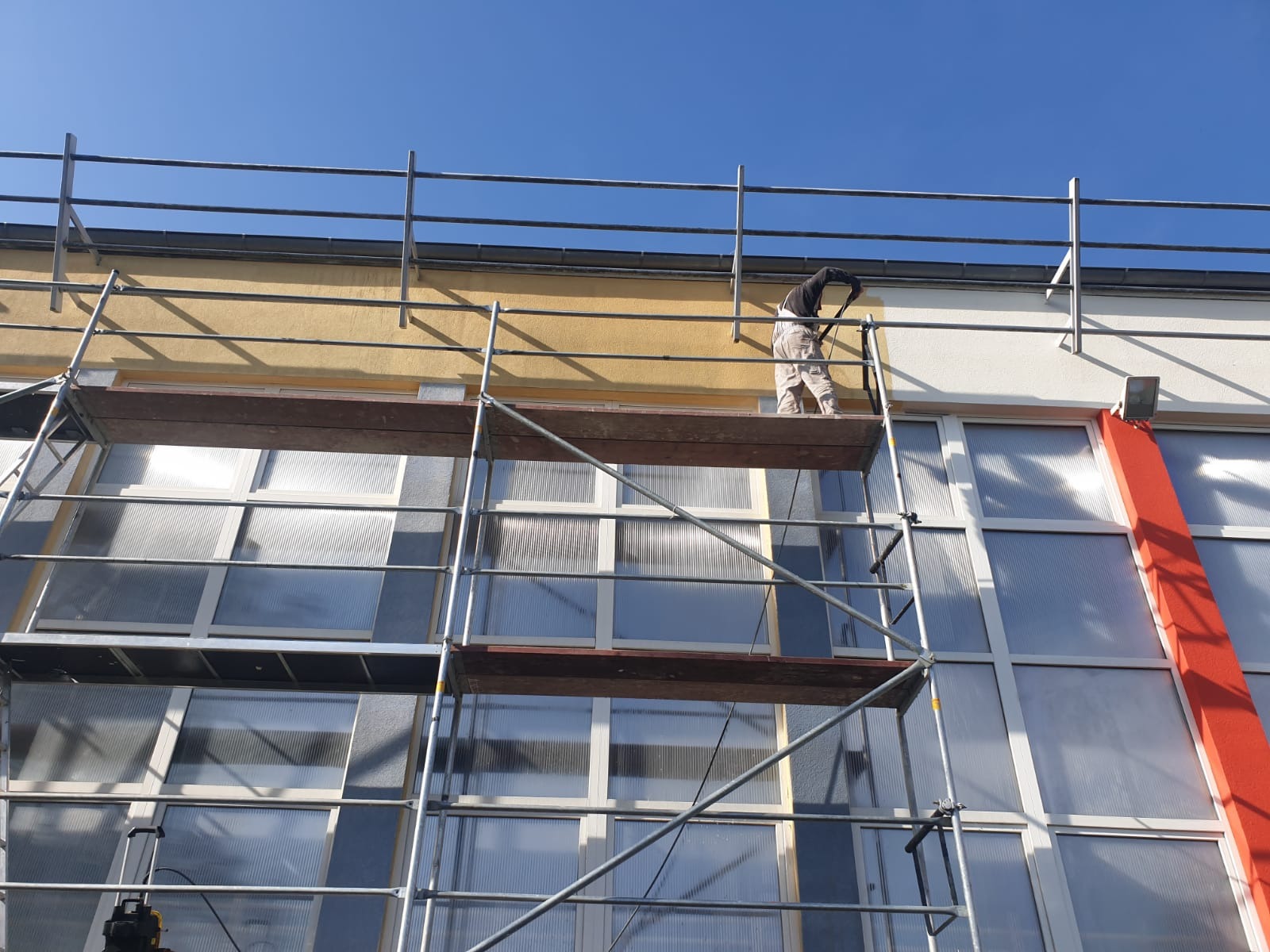 Pracownik na rusztowaniu maluje elewację budynku, widoczne okna z poliwęglanu, fragment pomarańczowej kolumny i niebieskie niebo.