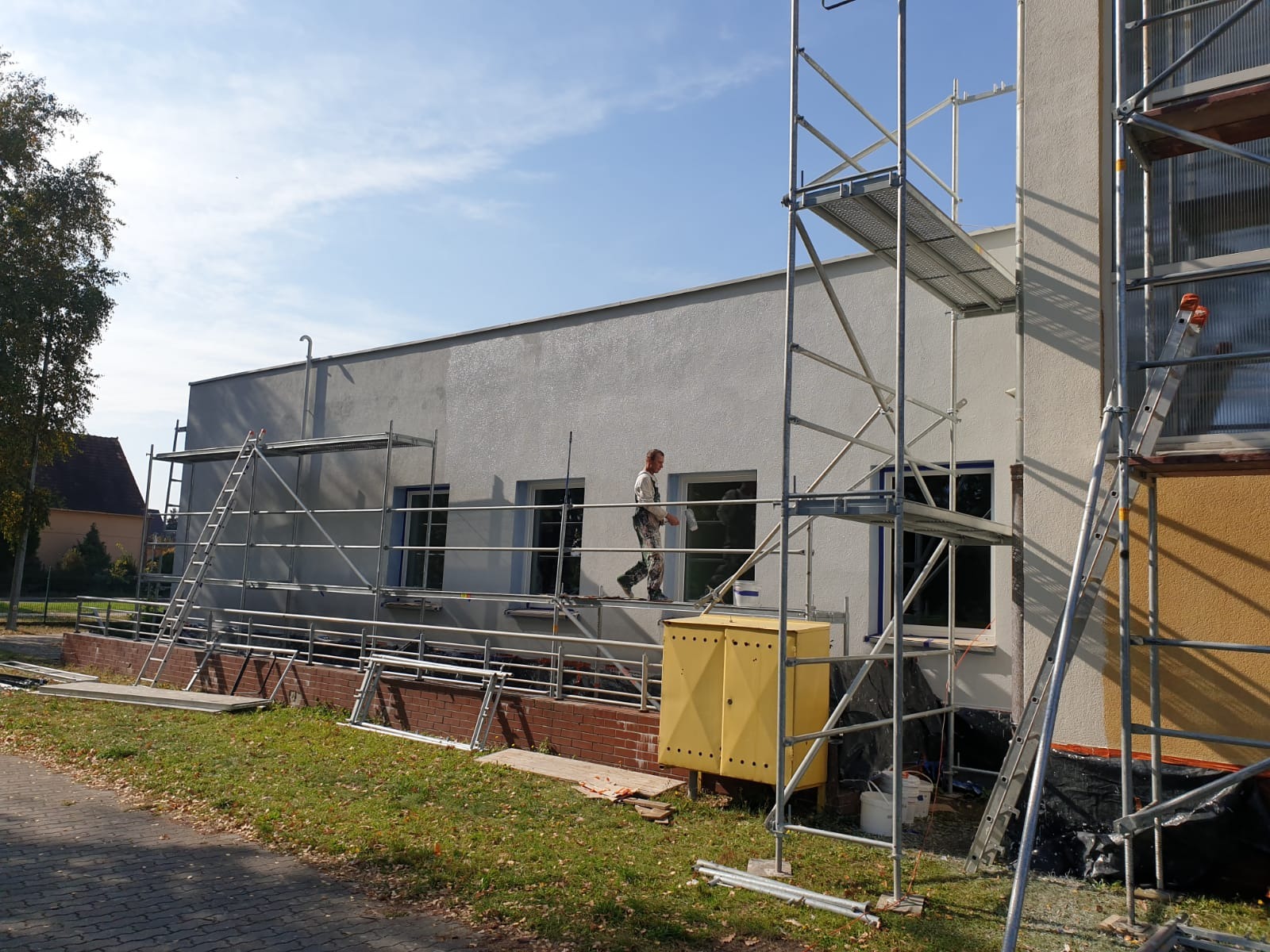 Pracownik w kombinezonie roboczym porusza się po rusztowaniu na tle świeżo otynkowanej, jasnoszarej elewacji budynku z ciemnymi oknami.