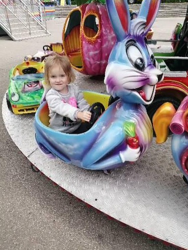 Dziewczynka siedzi w karuzeli w kształcie niebieskiego królika trzymającego marchewkę, w tle inne pojazdy karuzelowe.