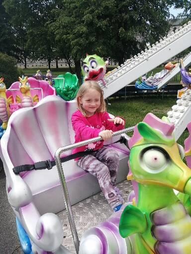 Dziewczynka w różowej bluzce siedzi w karuzeli w kształcie muszli z elementami morskimi, w tle widoczne inne karuzele i drzewa.