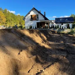 Przygotowany teren pod budowę domu jednorodzinnego, widoczne wykopane fundamenty, częściowo ogrodzony teren, w tle dom w stylu nowoczesnym z ciemnymi elementami wykończenia.