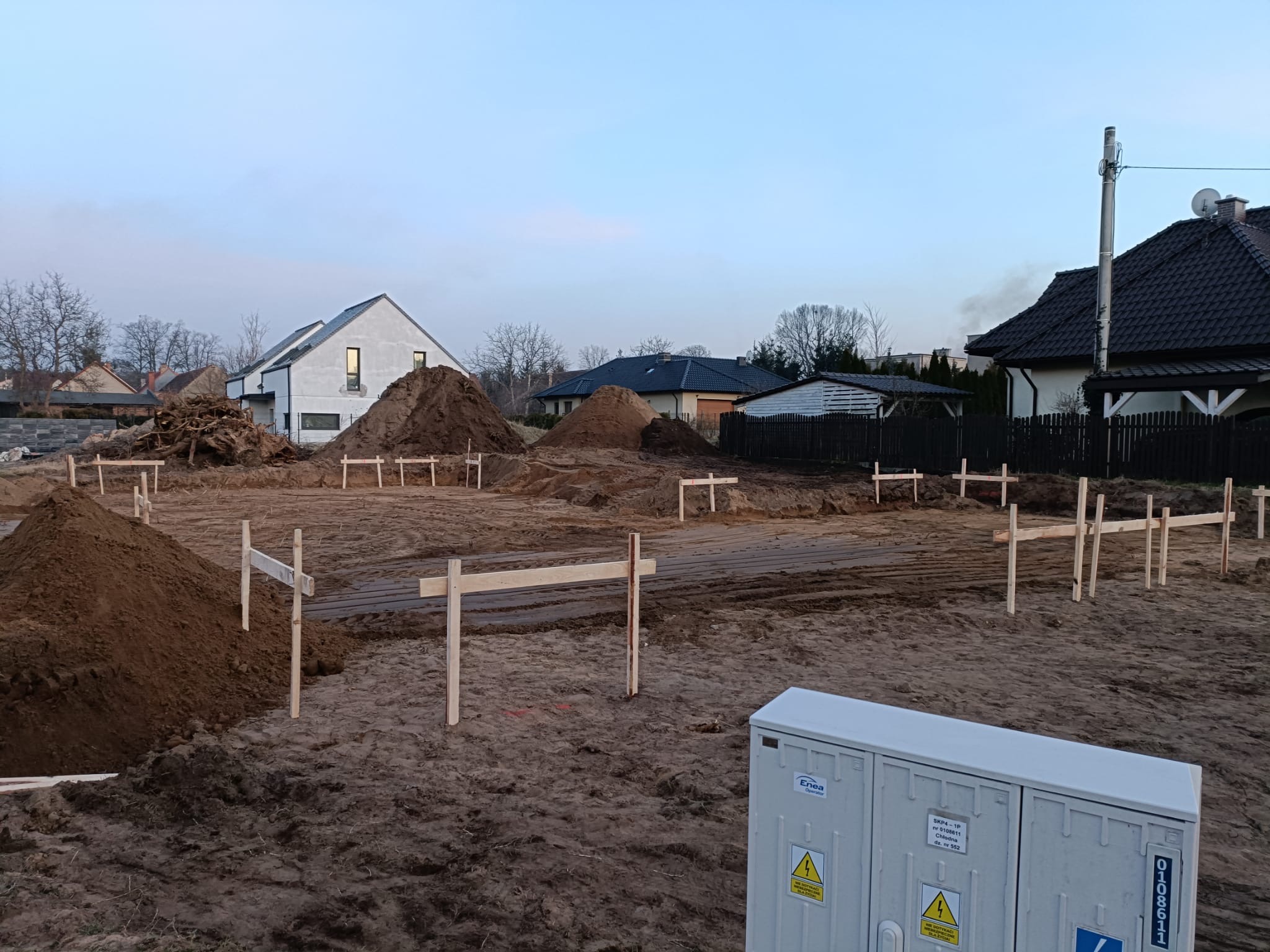Przygotowanie terenu pod budowę domu: wytyczone fundamenty z drewnianymi stemplami, pryzmy ziemi, skrzynka elektryczna Enea w rogu