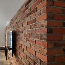 Ściana działowa z czerwonej cegły w nowoczesnym wnętrzu, z wnęką na telewizor, widoczny st&oacute;ł i krzesła w tle.